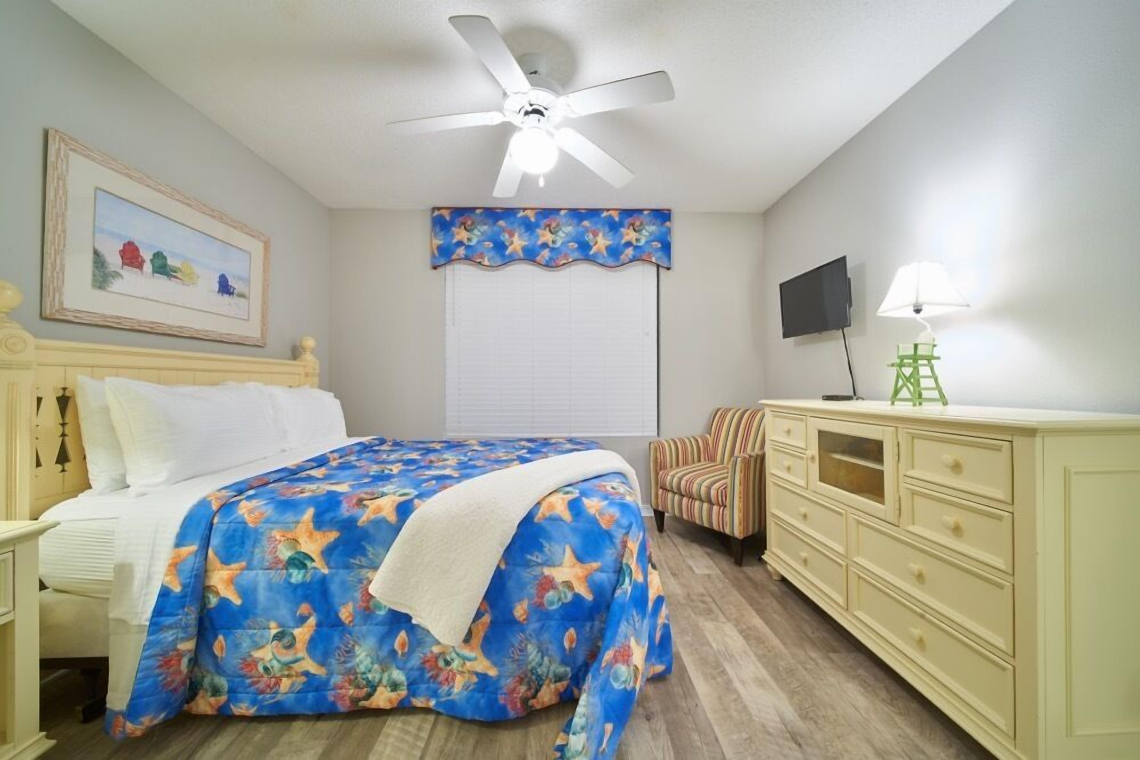 A bedroom with a bed, a ceiling fan, a TV, a lamp, and a dresser.