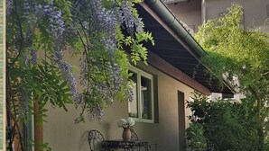 Un jardin avec une table et des chaises, entouré d’une végétation luxuriante et de fleurs violettes.