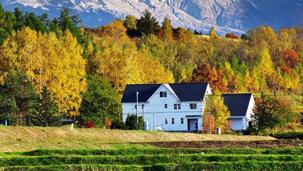 Une maison blanche avec un toit sombre, entourée d’arbres aux couleurs automnales et, au loin, d’une montagne dont le sommet est enneigé.