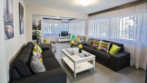 A modern living room with two sofas, a coffee table, and a TV. There are curtains on the windows and a plant on the windowsill.