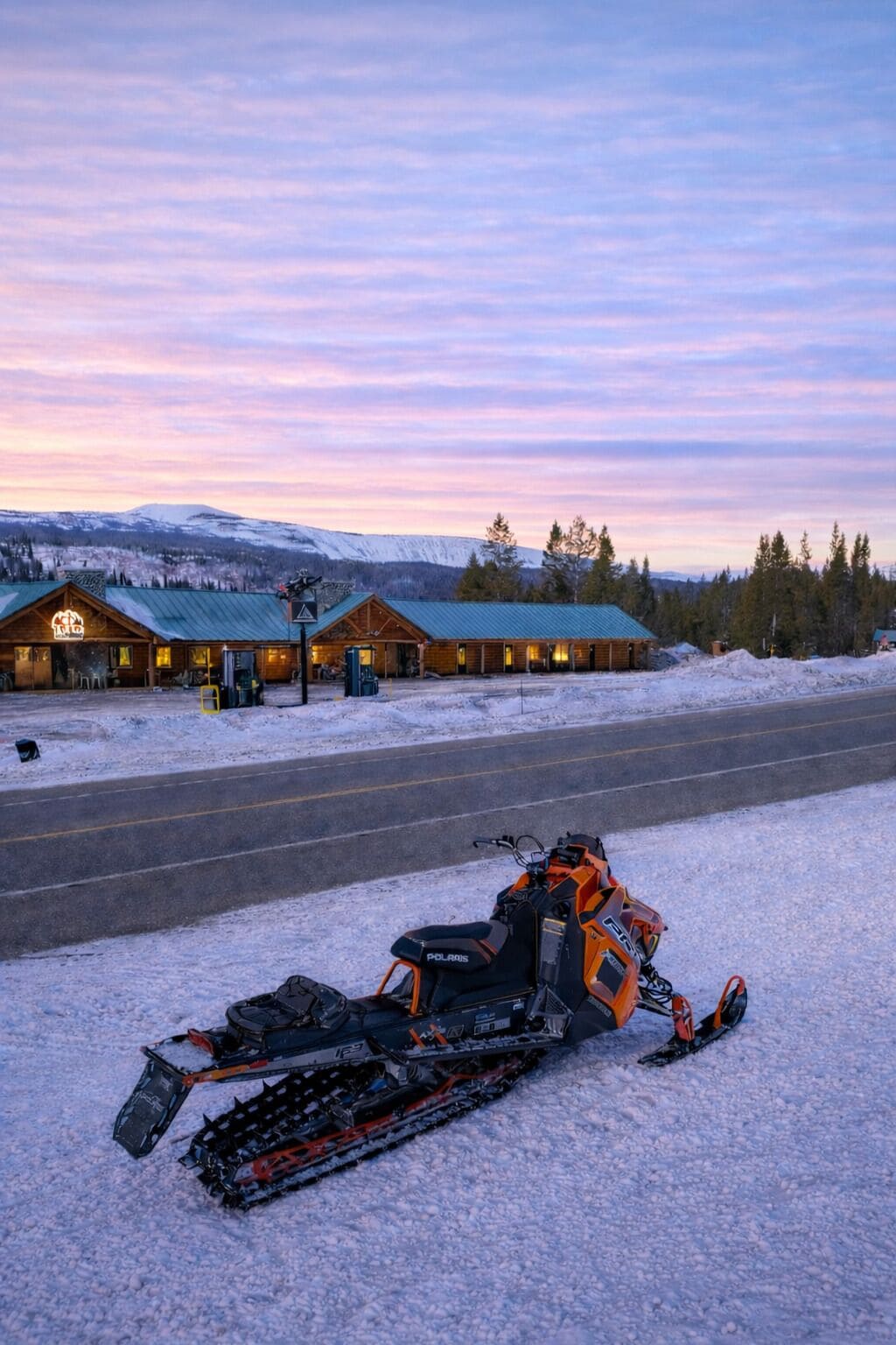 Una moto de nieve estacionada frente a un albergue, con una carretera y un paisaje nevado al fondo.
