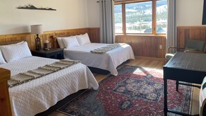 A room with two beds, a table, a chair, a lamp, and a view of a snowy landscape through large windows. - Lava Mountain Lodge (Dubois)