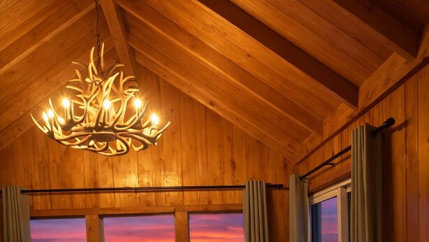 A wooden interior with a chandelier, a view of a sunset through windows, a rustic chair, a hat, and a suitcase. - Lava Mountain Lodge (Dubois)