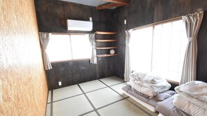 Une pièce avec un sol recouvert de tatamis, un futon servant de lit, une étagère ornée d’objets décoratifs et une fenêtre avec des rideaux.
