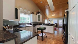 A modern kitchen with a wooden ceiling, white cabinets, a central island, and a dining area with a view.