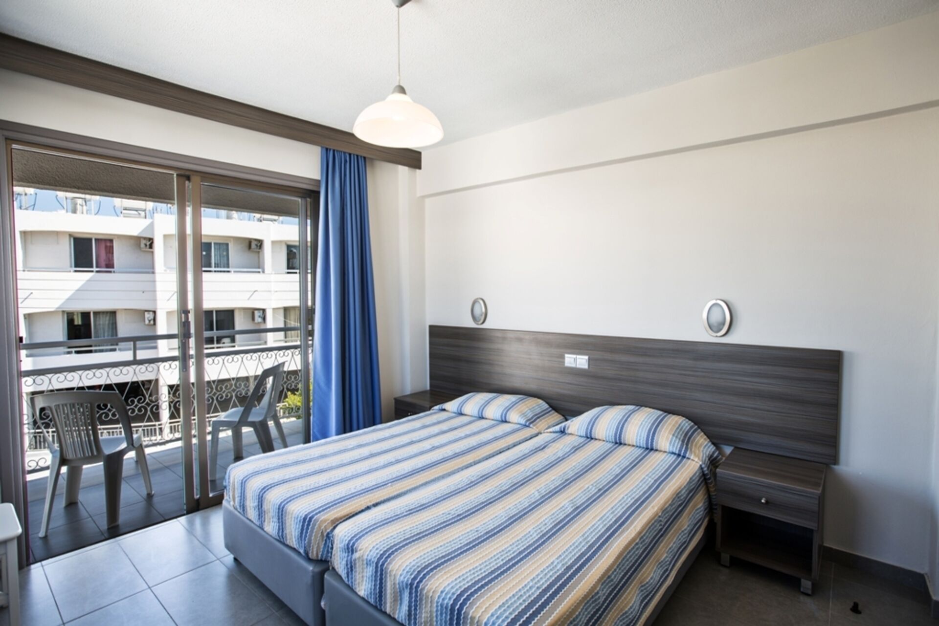 Ein Hotelzimmer mit Bett, Nachttischen, einem Fenster mit Blick auf Gebäude und einem Balkon mit Stühlen.
