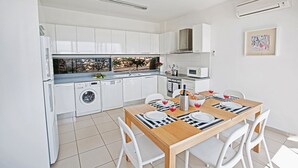 Une cuisine moderne avec des meubles blancs, un lave-linge encastré, un coin repas avec une table en bois et des chaises blanches, et une vue sur un jardin par la fenêtre.