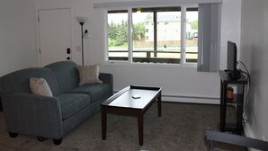 A living room with a grey sofa, a small table, a TV stand, and a window with a view of a fence and buildings. - Corner Midtown Apartment (Anchorage)