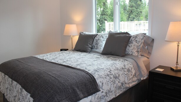 A bedroom with a bed, two bedside tables with lamps, a window with a view of trees, and a white wall. - Corner Midtown Apartment (Anchorage)