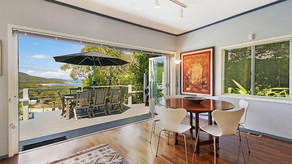Uma área de refeições moderna com mesa e cadeiras de madeira, um guarda-sol grande e vista para o exterior.