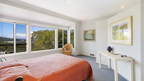 Quarto com cama grande, uma cadeira, escrivaninha e vista para o exterior através de amplas janelas.