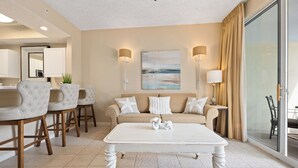 Un salon moderne avec une table basse blanche, un canapé beige et des fauteuils assortis. On y trouve deux lampes fixées au mur, un tableau et une porte-fenêtre coulissante qui donne sur un balcon.