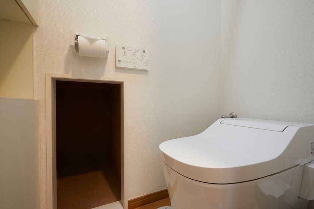 A small, clean bathroom with a toilet, a roll of toilet paper, and a power outlet on the wall.