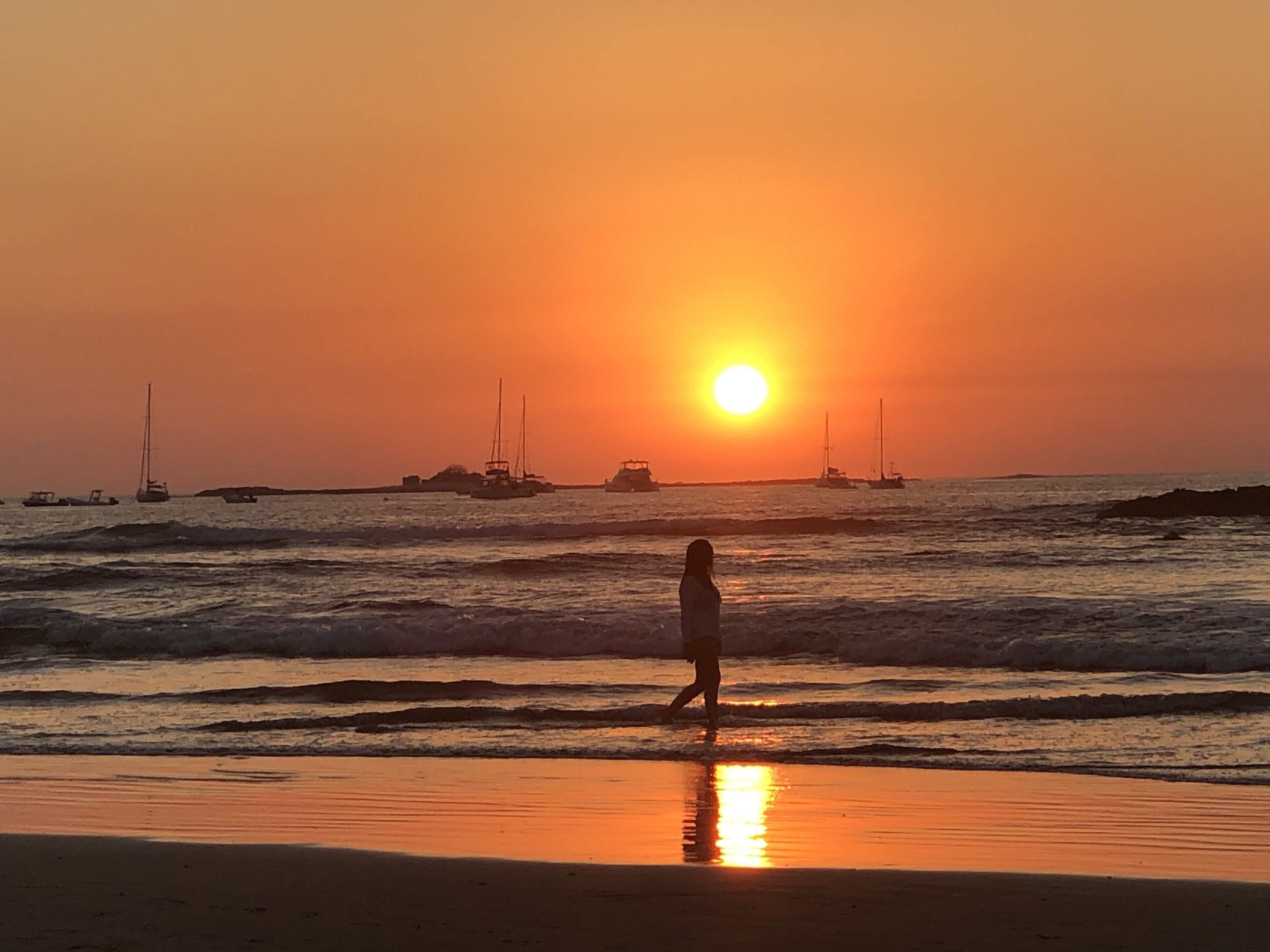 Eine Strandansicht mit einer Person, die am Ufer entlangspaziert, Booten im Wasser und einem Sonnenuntergang im Hintergrund.