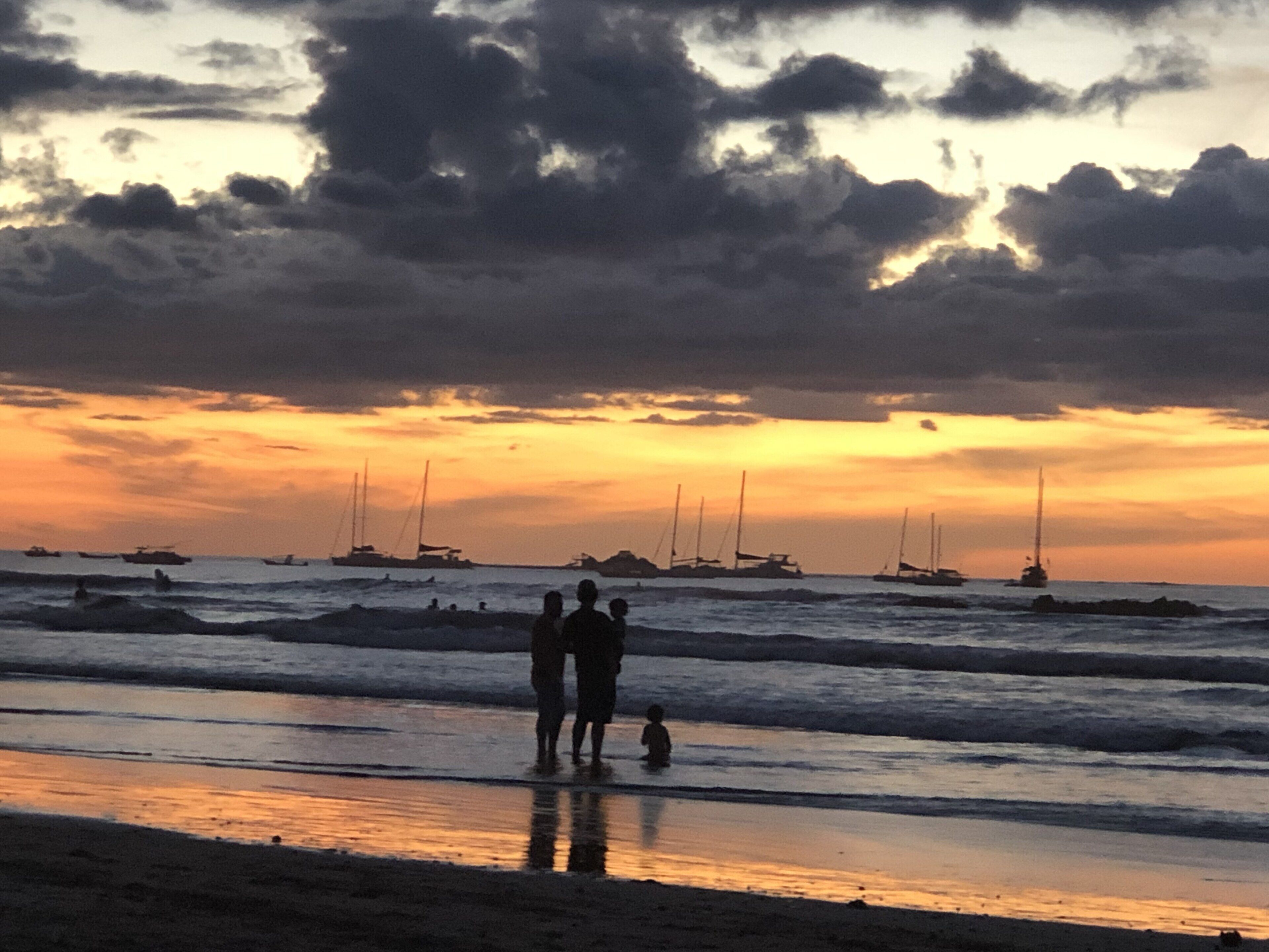 A beach scene with people standing by the water, boats in the distance, and a sunset sky.