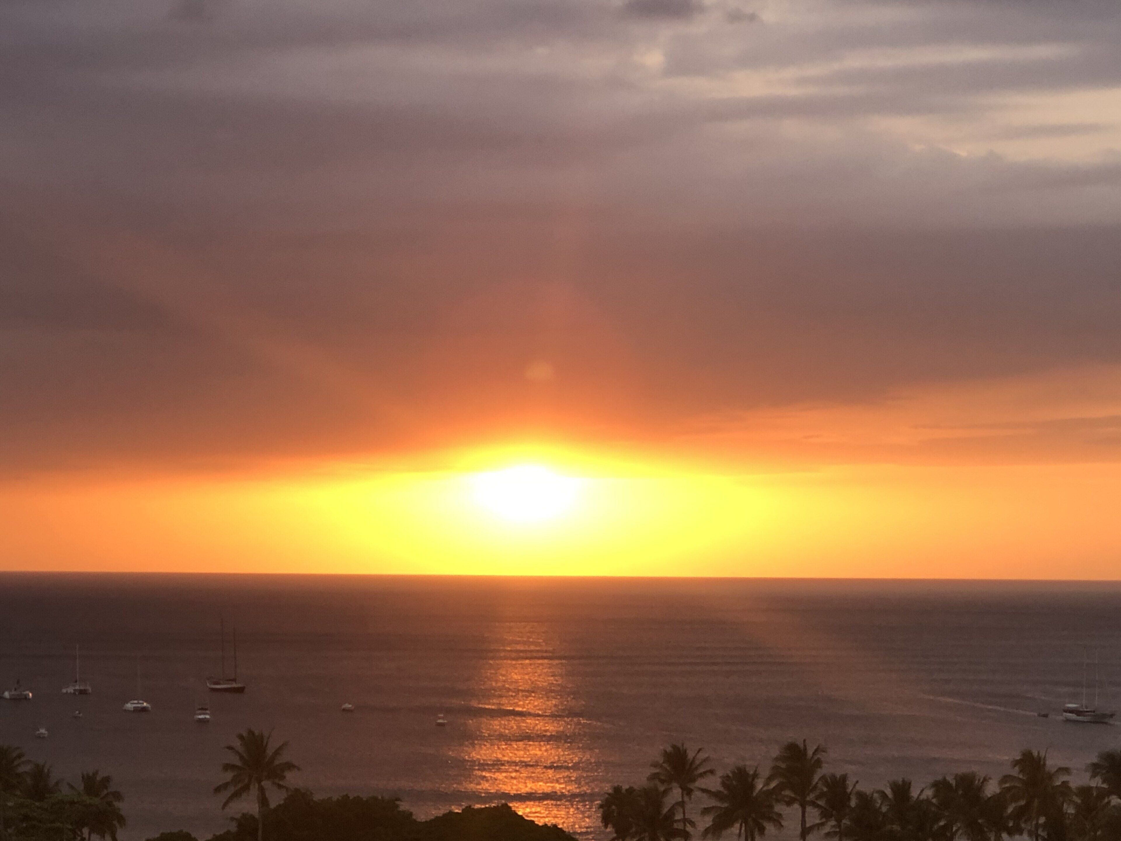 A sunset over a calm sea with boats and palm trees.