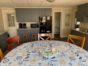 A kitchen with grey cabinets, a black refrigerator, a microwave, and a coffee maker. There are two wooden stools and a table with a nautical-themed tablecloth.