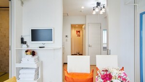 Ett hotellrum med en tv, en hylla med handdukar, en orangefärgad stol och en blomsteruppsättning.