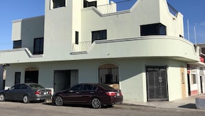 Ein modernes, zweistöckiges Gebäude mit Flachdach und Balkon. Vorne sind zwei Autos geparkt, und ein verschlossener Eingang ist sichtbar.