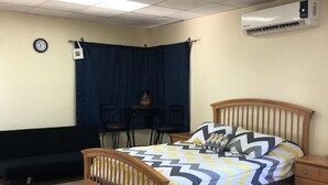 Ein Schlafzimmer mit einem Bett, einem hölzernen Kopfteil, einem Nachttisch, einem kleinen Tisch mit einer Vase, einem schwarzen Sofa, einem blauen Vorhang, einer Wanduhr und einer Klimaanlage.
