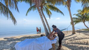 Sesión de masoterapia en una playa arenosa con palmeras y una hamaca.