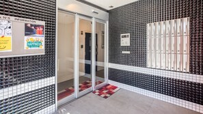 Een hotellobby met een glazen deur, een betegelde muur met genummerde kluisjes en een welkomstbord.