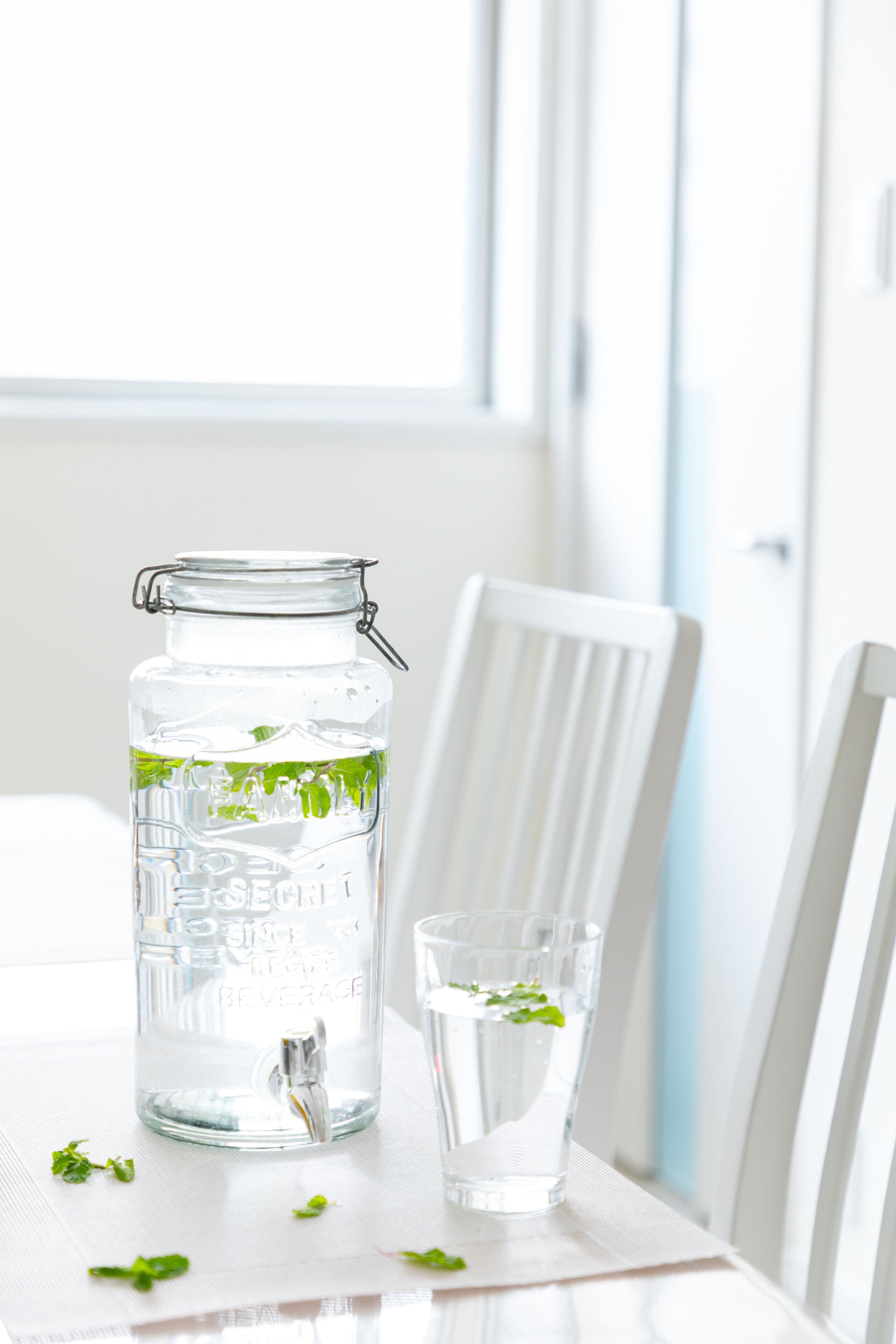 A clear glass jar with a metal clasp and a metal spout, filled with water and mint leaves, placed on a white table next to a glass of water.