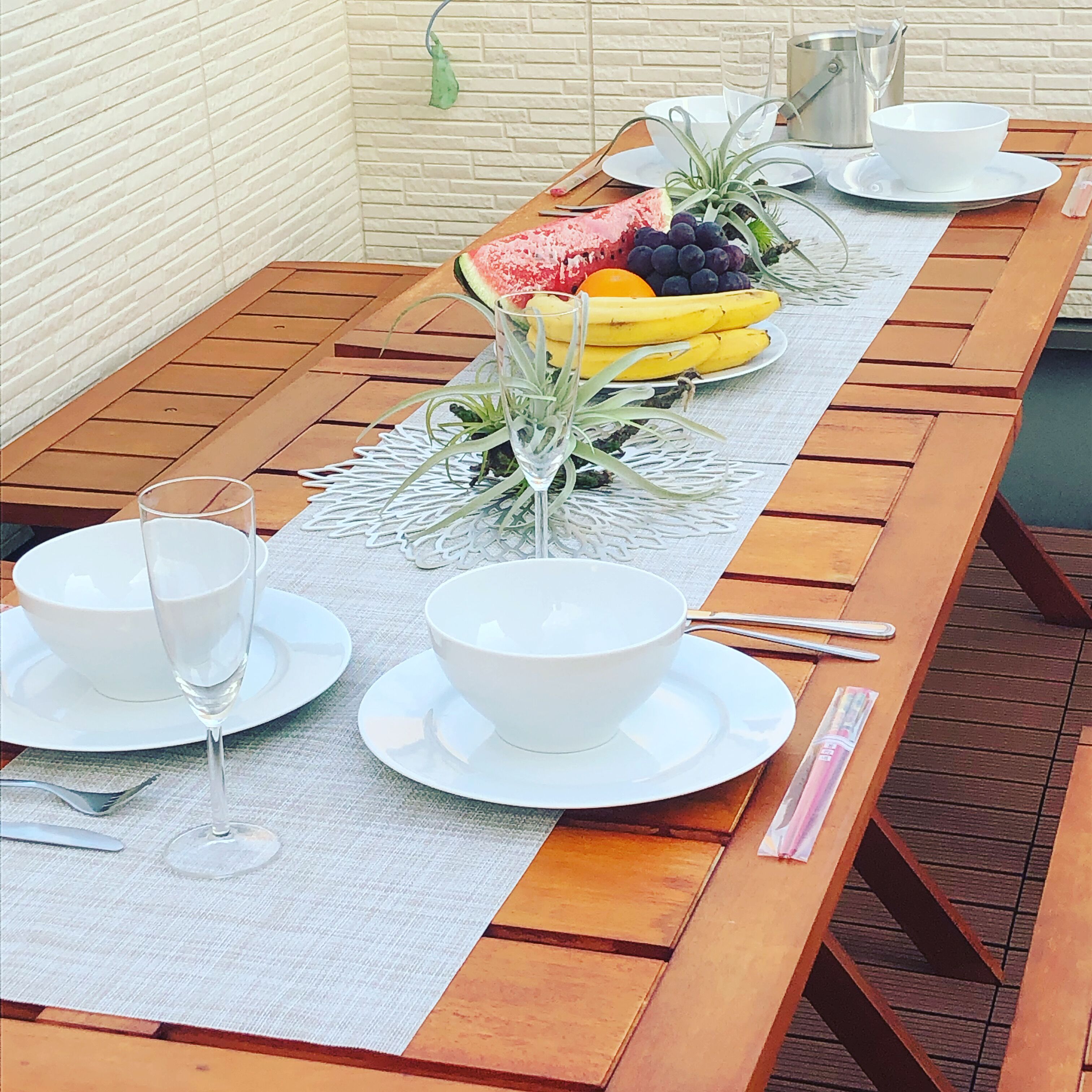 A wooden dining table with white plates, bowls, and a glass of water. There is a fruit arrangement on the table, including bananas, grapes, and watermelon.