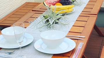 A wooden dining table with white plates, bowls, and a glass of water. There is a fruit arrangement on the table, including bananas, grapes, and watermelon.