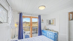 A bedroom with a bed, a dresser, a window with curtains, a table and chairs outside, and a view of the landscape.