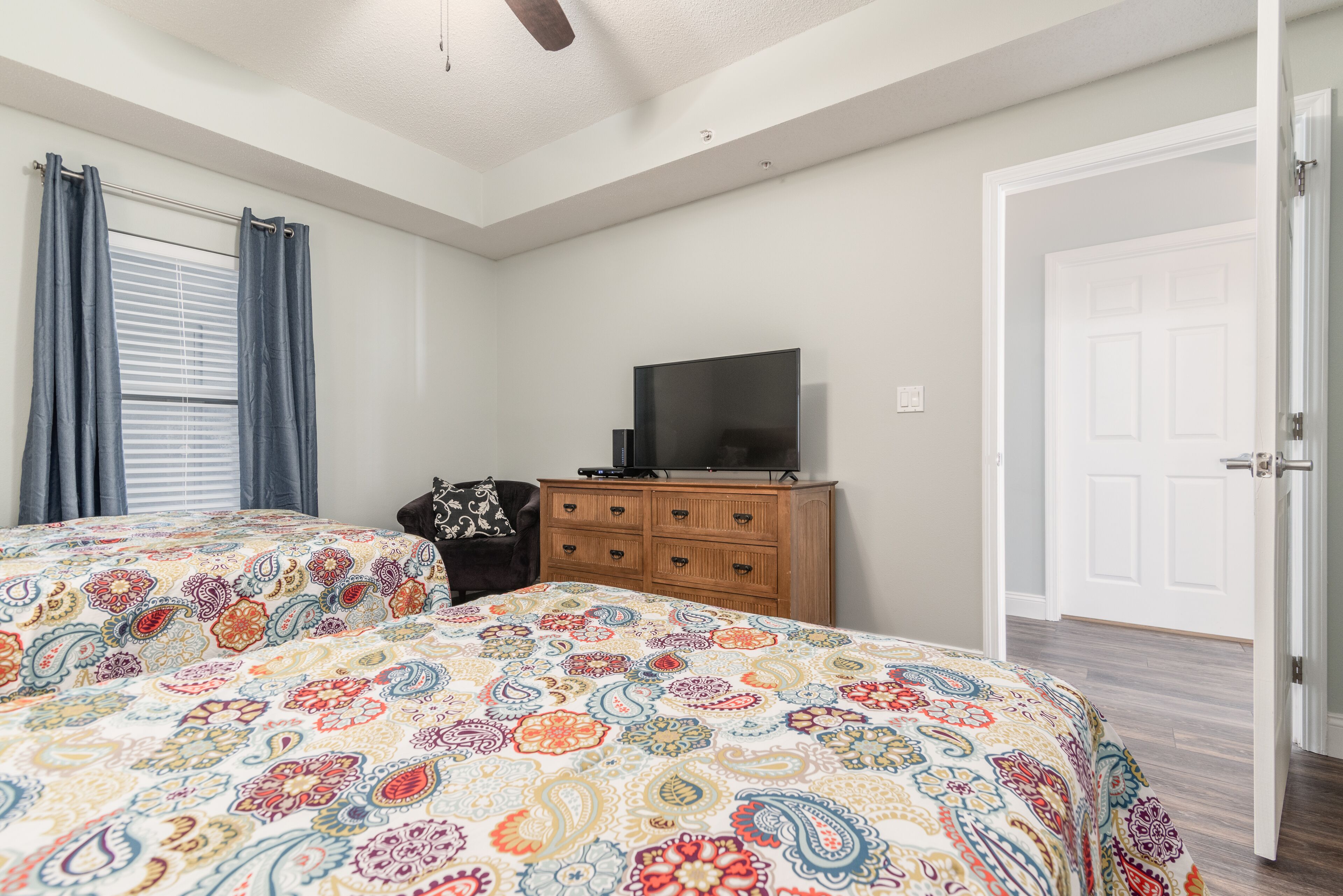 Une chambre à coucher avec un grand lit, un téléviseur posé sur une commode en bois, une porte et une fenêtre avec des rideaux.
