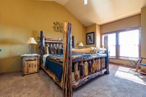 A bedroom with a rustic bed frame, a small wooden nightstand, a chair, and a large window with blinds.