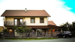 Ein zweistöckiges Haus mit Balkon, einem geparkten Auto davor und einem Zaun mit blühenden Pflanzen.