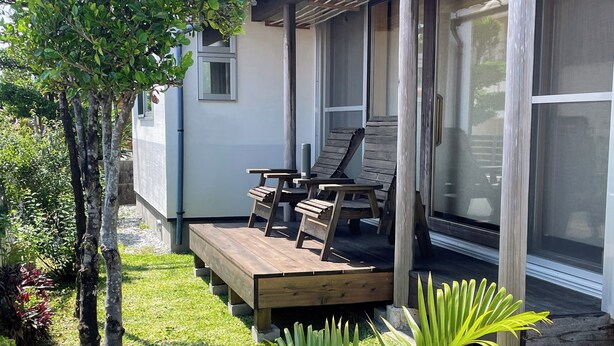 Eine Holzterrasse mit Gartenmöbeln, umgeben von üppigem Grün und einem Gebäude mit großen Fenstern.