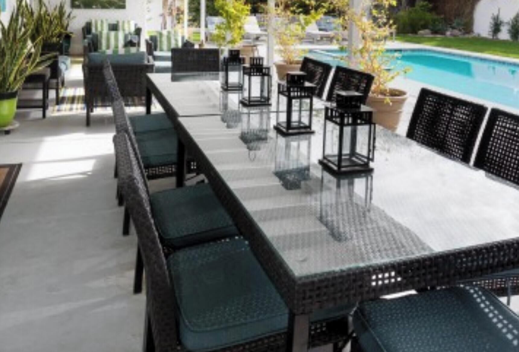 A poolside area with a long table and chairs, surrounded by potted plants and a view of a swimming pool.