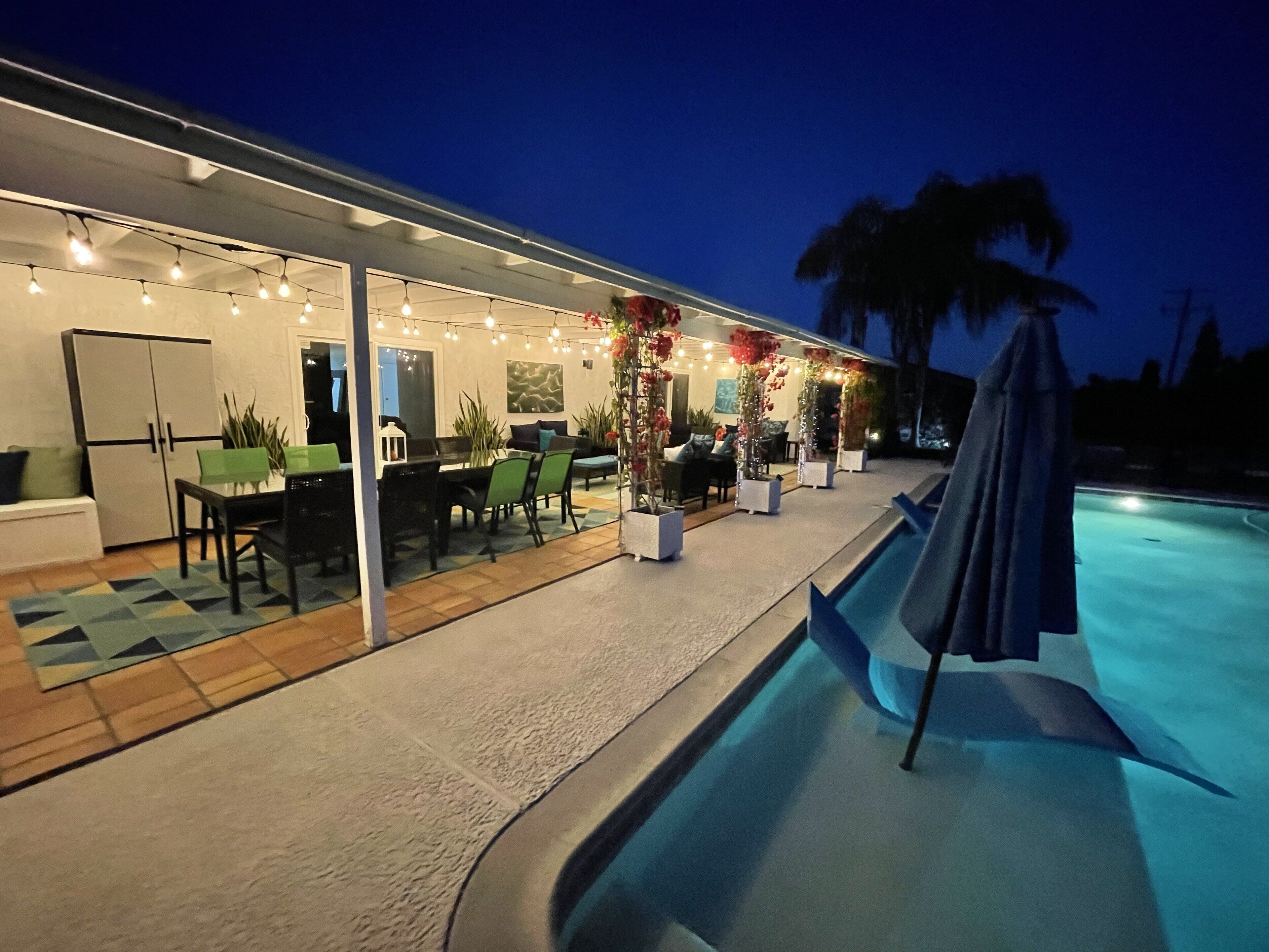 Un área de piscina al aire libre con un porche cubierto, mobiliario y luces colgantes.
