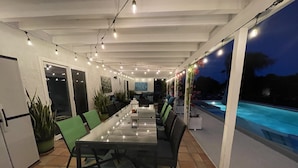 Een eetgedeelte met een lange tafel, stoelen en sfeerverlichting. Er is een zwembad zichtbaar door de glazen wand.