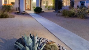 Een woonhuis met een verharde oprit naar een voordeur, omgeven door woeste planten en een berg op de achtergrond.