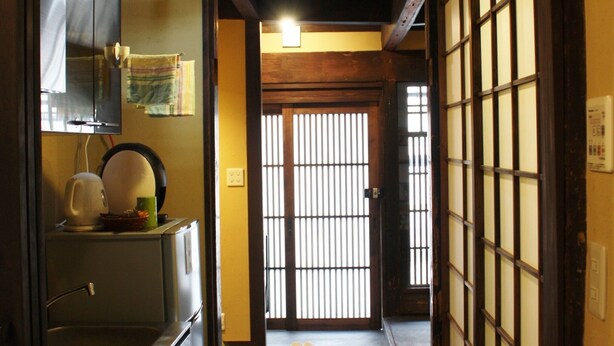 Ein schmaler Flur mit einer traditionellen japanischen Schiebetür, einer Deckenleuchte und einem Kühlschrank mit Wasserkocher darauf.