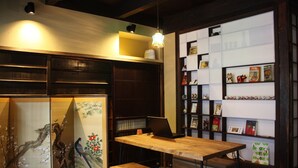 Ein traditioneller japanischer Raum mit Tatami-Böden, einem niedrigen Holztisch und einem Bücherregal mit Büchern und dekorativen Gegenständen.