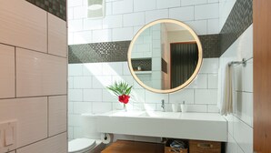Une salle de bain moderne avec un lavabo blanc, un miroir rond et des toilettes. On y trouve des meubles en bois sous le lavabo et une petite plante posée sur le comptoir.