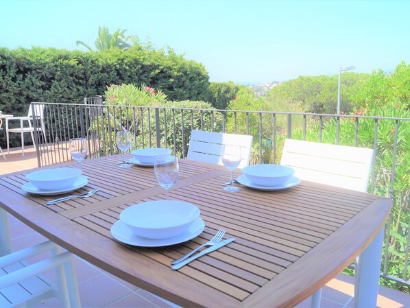 An outdoor dining area with a wooden table set for four, surrounded by white chairs, overlooking a lush garden and a distant view.