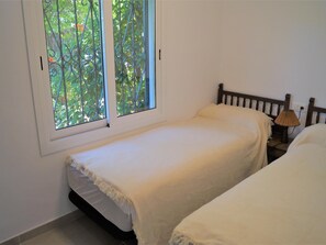 A bedroom with two single beds, a window with a metal grill, and a bedside lamp.