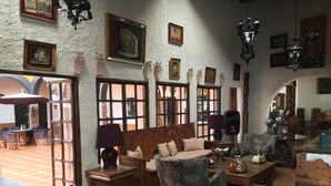 Ein gemütliches Interieur mit einer Couch, Sesseln und einem Esstisch. An den Wänden hängen Bilder in Rahmen und eine Kristallleuchter ist angebracht.