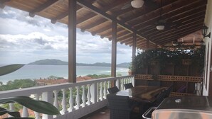 Ein überdachter Außenbereich mit Tisch, Stühlen, einem Grill und Blick auf das Meer und die fernen Hügel.