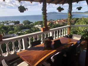 Mesa de madeira com cadeiras numa varanda com vista para a costa, plantas penduradas e balaústres brancos.