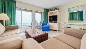 A living room with beige sofas, a brown ottoman, a blue armchair, and a TV on a white cabinet. - Crystal Sands #101B * Beach Chair Set Included * Salt Water Vacations (Destin)
