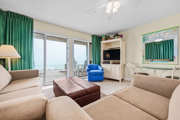 A living room with beige sofas, a brown ottoman, a blue armchair, and a TV on a white cabinet. - Crystal Sands #101B * Beach Chair Set Included * DON'T MISS FALL BREAK! (Destin)