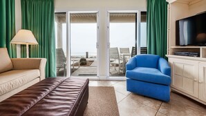 A living room with a sofa, a blue chair, a TV, and a view of the beach through the sliding doors. - Crystal Sands #101B * Beach Chair Set Included * Salt Water Vacations (Destin)