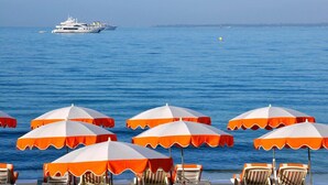 Une plage bordée de rangées de parasols orange et blancs et de chaises longues, avec un yacht en arrière-plan et une eau bleue et calme.